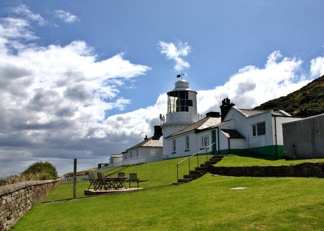 A lighthouse with a white tower and a green base, situated on a grassy hill with a stone wall and a picnic table.