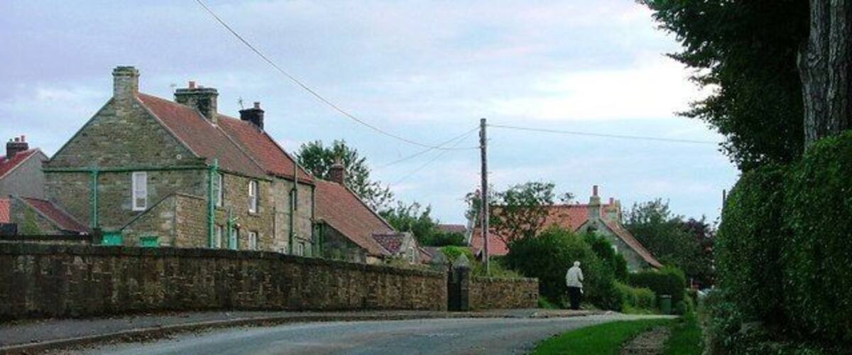 A rural road with stone walls, houses with red roofs, and a grassy area.