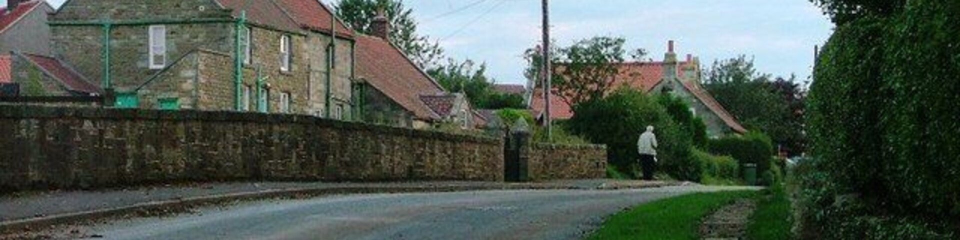 A rural road with stone walls, houses with red roofs, and a grassy area.