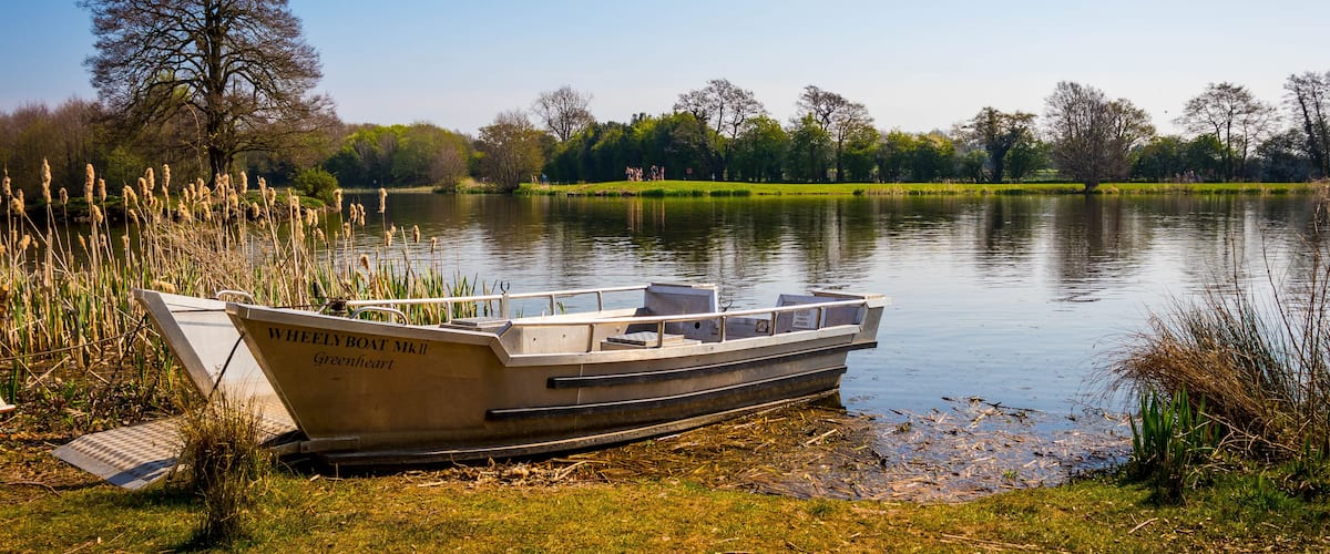 A boat named "Wheely Boat MkII" docked at a lakeside with grassy banks and trees in the background.