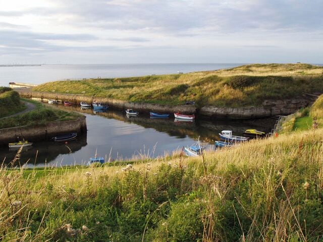 A coastal landscape with boats docked in a narrow waterway, surrounded by grassy hills.