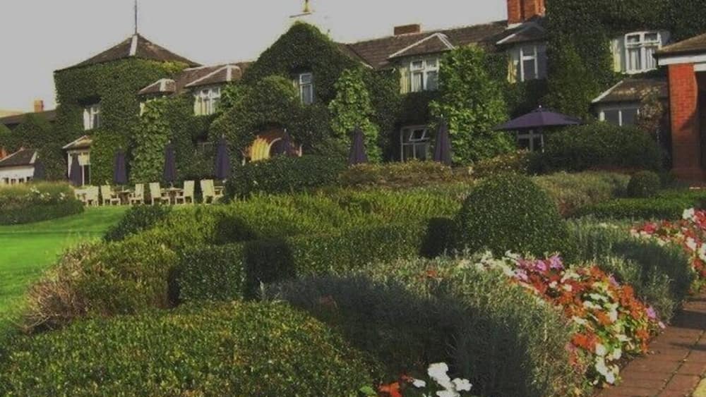 A large house with ivy-covered walls, a garden with various flowers, and a clear sky.
