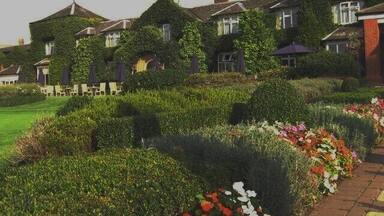 Een groot huis met muurranken, een tuin met verschillende bloemen en een heldere lucht.