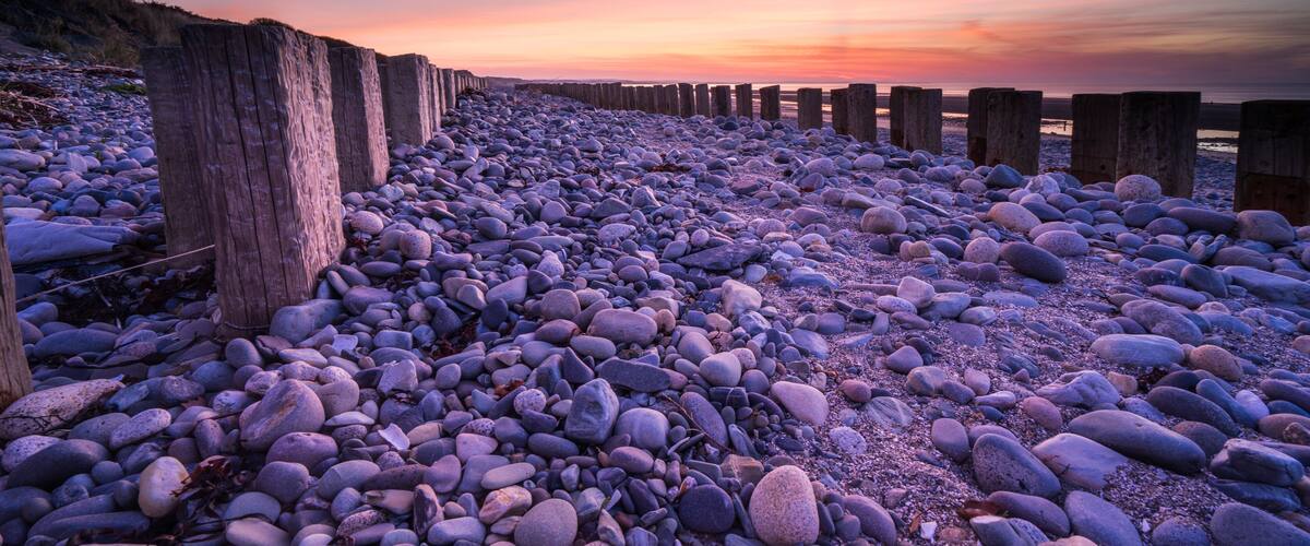 A pebbled beach with wooden posts and a sunset sky.