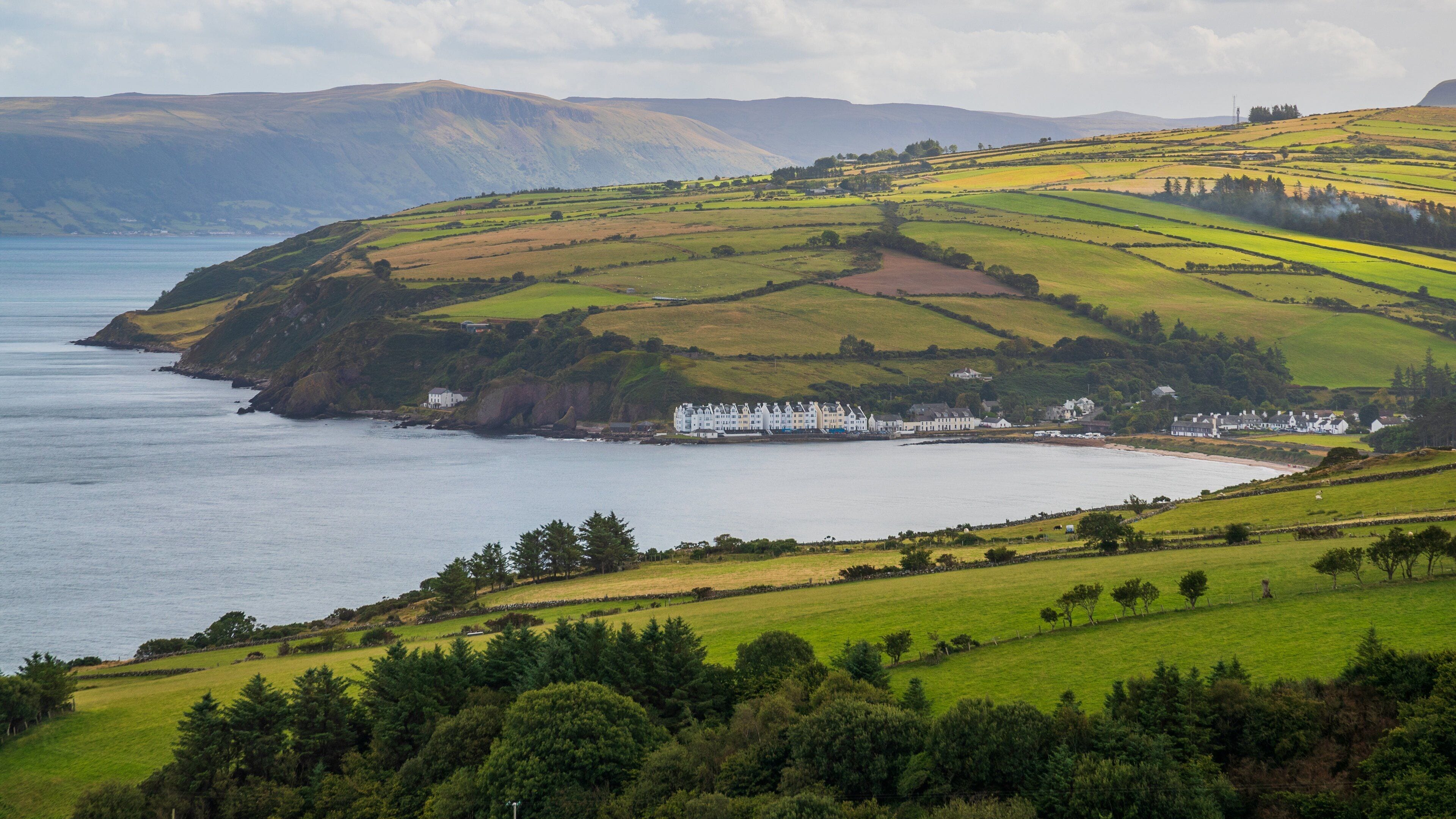 A coastal landscape with rolling hills, a body of water, and a row of white buildings along the shore.