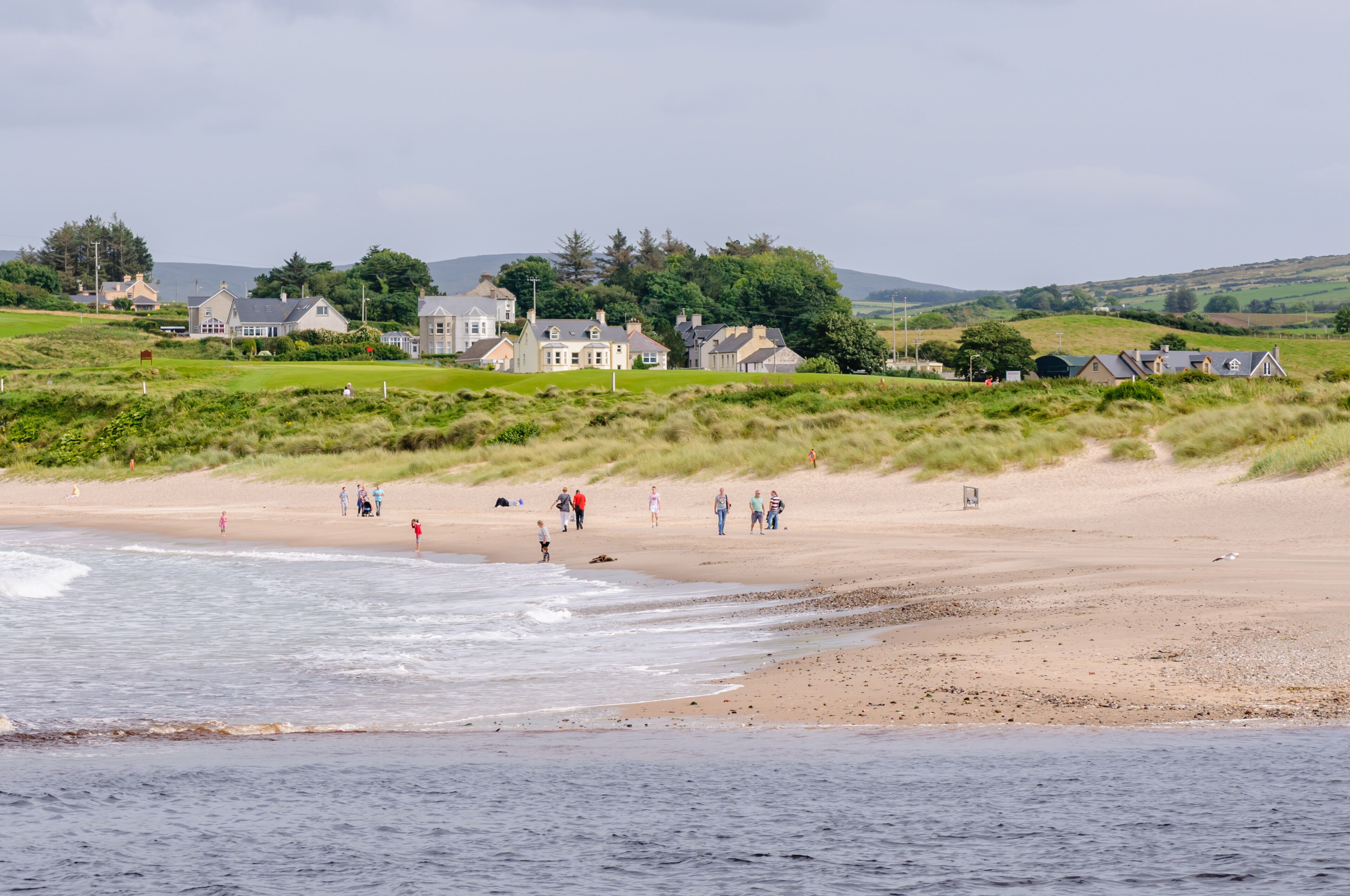 Una playa costera con gente caminando, una orilla arenosa y casas al fondo.