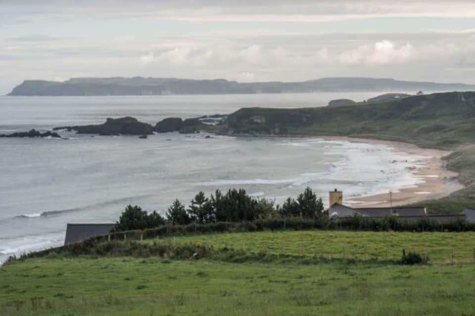 A coastal landscape with a grassy foreground, a sandy beach, rocky outcrops, and a distant shoreline.