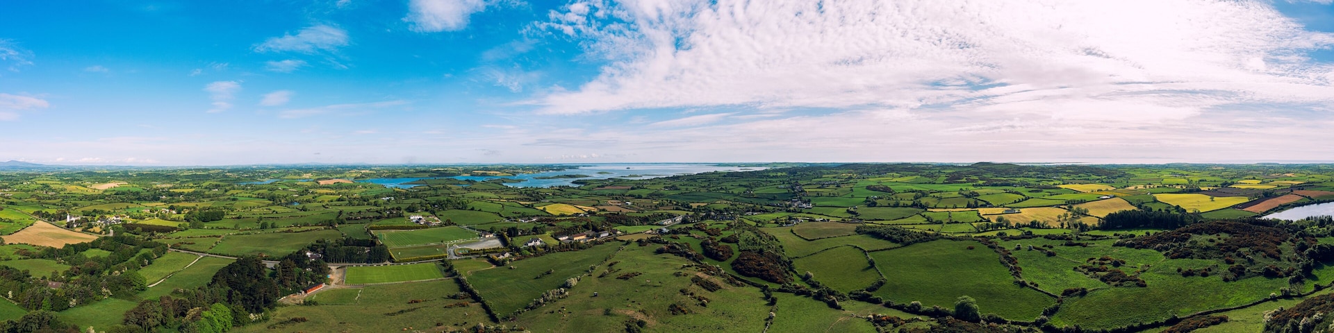 Vista panorâmica de uma paisagem rural com campos verdes ondulados, casas espalhadas e um lago ao longe.