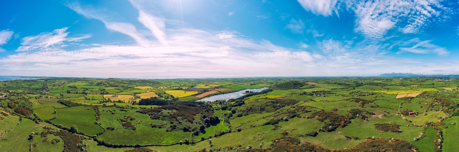 Una vista panorámica de un valle verde exuberante con campos, un río y montañas distantes bajo un cielo parcialmente nublado.