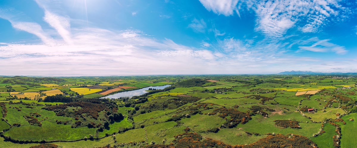 A panoramic view of a lush green valley with fields, a river, and distant mountains under a partly cloudy sky.