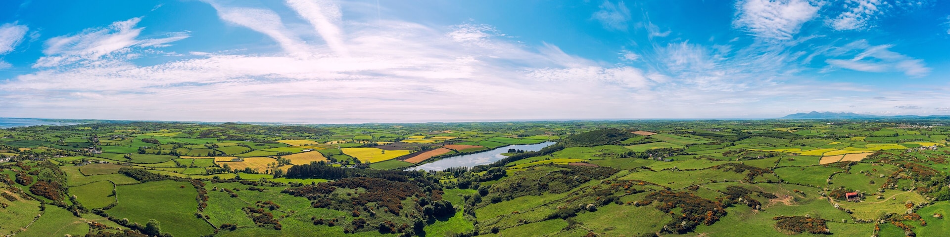 A panoramic view of a lush green valley with fields, a river, and distant mountains under a partly cloudy sky.