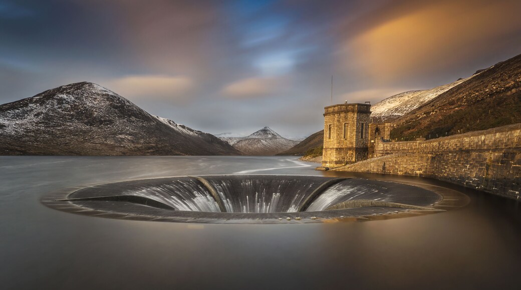 View of the bell-mouth spillway and stone tower at Silent Valley Reservoir with snow-dusted mountains under a sunset sky in Newry, Northern Ireland, United Kingdom.