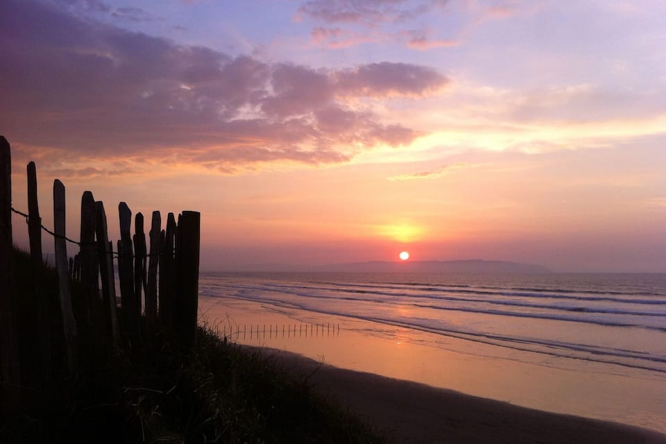 Een zonsondergang boven een strand, met een houten omheining op de voorgrond.