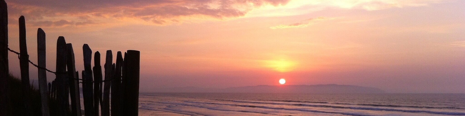 Um pôr do sol sobre uma praia, com uma cerca de madeira ao primeiro plano.
