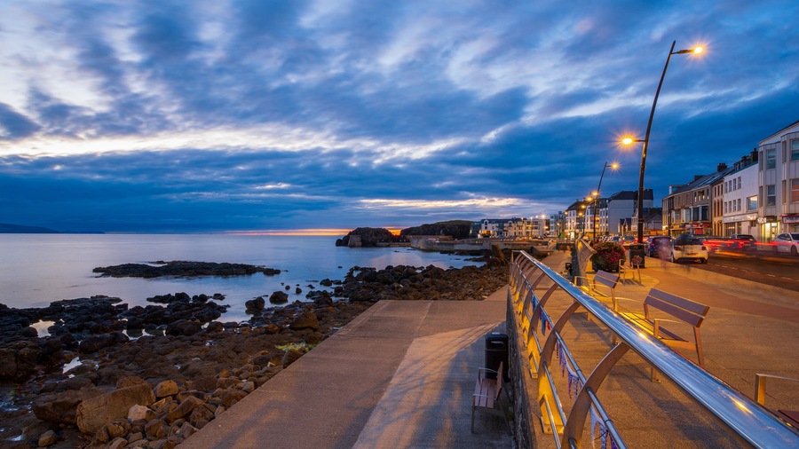 A coastal promenade at dusk with a rocky shoreline, a calm sea, and a row of buildings along the promenade.