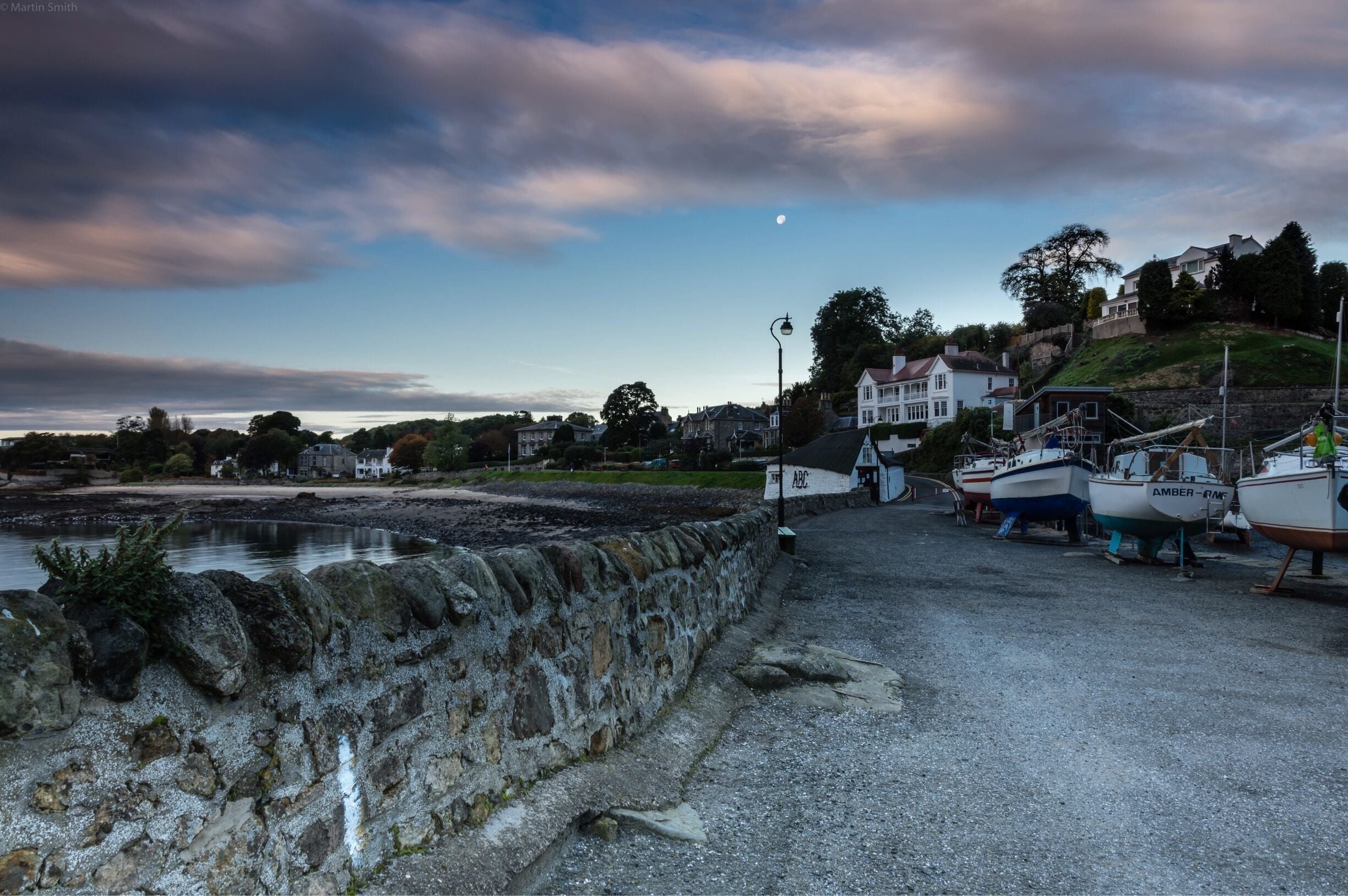 A coastal town with stone walls, boats, and houses on a hill.