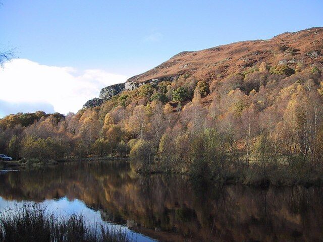 A calm lake reflecting trees and a rocky hillside.