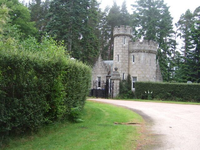A stone castle with a round tower, surrounded by lush greenery and a paved driveway.