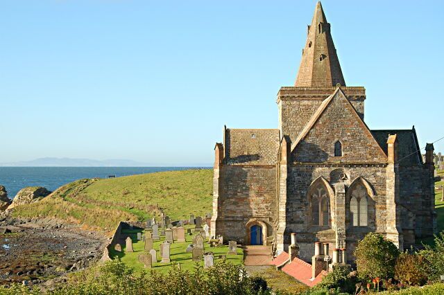Una iglesia de piedra histórica con una aguja alta, rodeada por un cementerio y con vistas al paisaje costero.