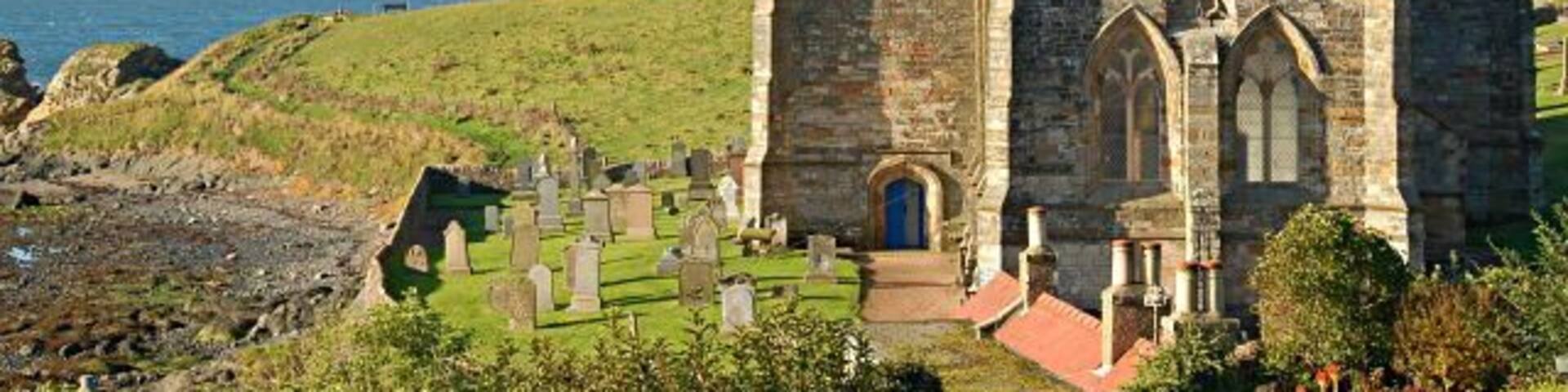 A historic stone church with a tall spire, surrounded by a graveyard and overlooking a coastal landscape.