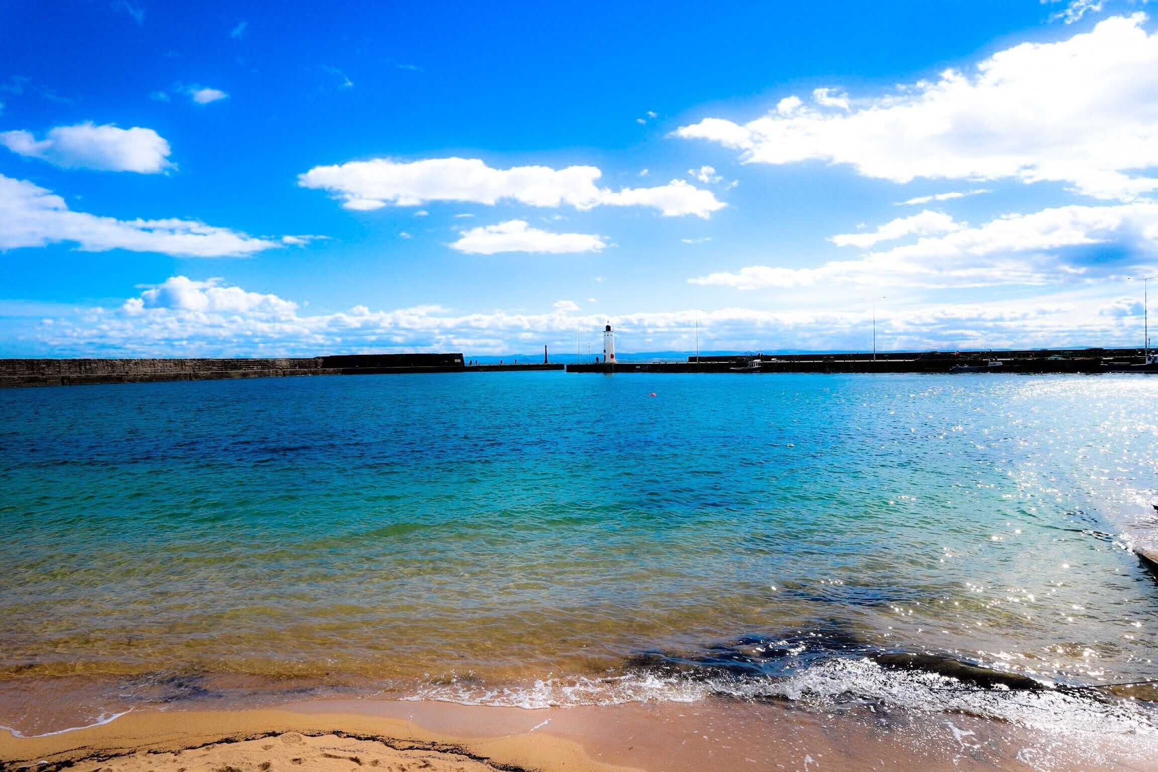 A beach with clear turquoise water, a sandy shore, and a lighthouse in the distance.
