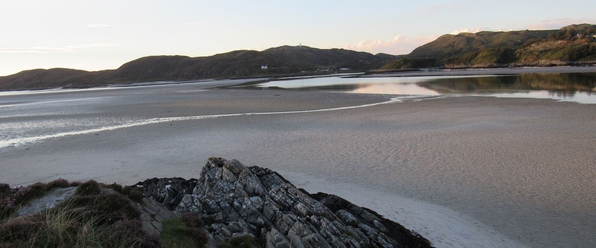 A coastal landscape with a rocky shoreline, calm water, and distant hills.