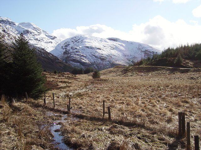 Paisagem montanhosa com picos nevados, um campo gramado e um pequeno riacho.