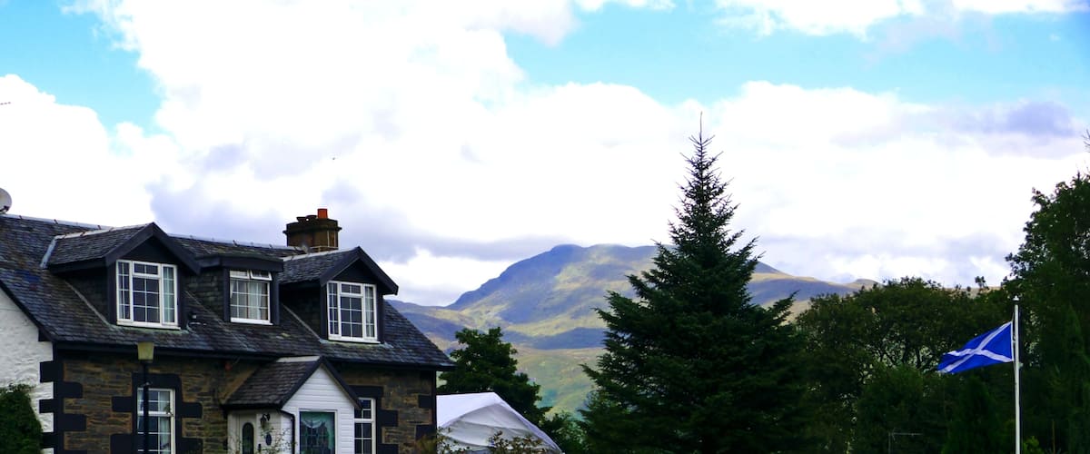 A stone house with a white front, a flag, and a mountain in the background.
