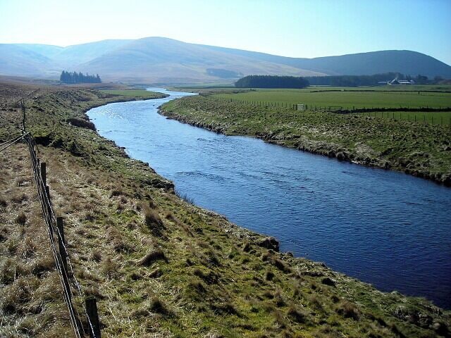 Ein Fluss schlängelt sich durch eine grüne Landschaft mit einem Zaun und fernen Bergen.