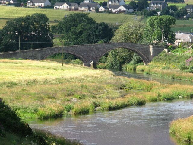 A stone bridge over a river, surrounded by grassy fields and houses in the background.