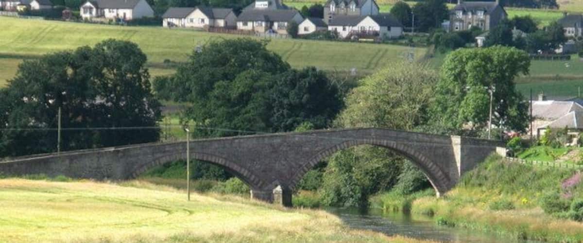 A stone bridge over a river, surrounded by grassy fields and houses in the background.