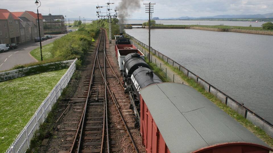 A train traveling along tracks next to a river, with buildings and a bridge in the background.