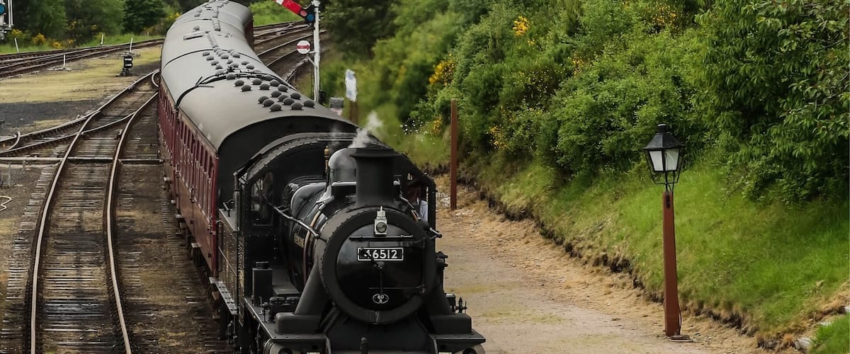 Une locomotive à vapeur sur les voies ferrées, entourée de verdure, avec un ciel nuageux en arrière-plan.