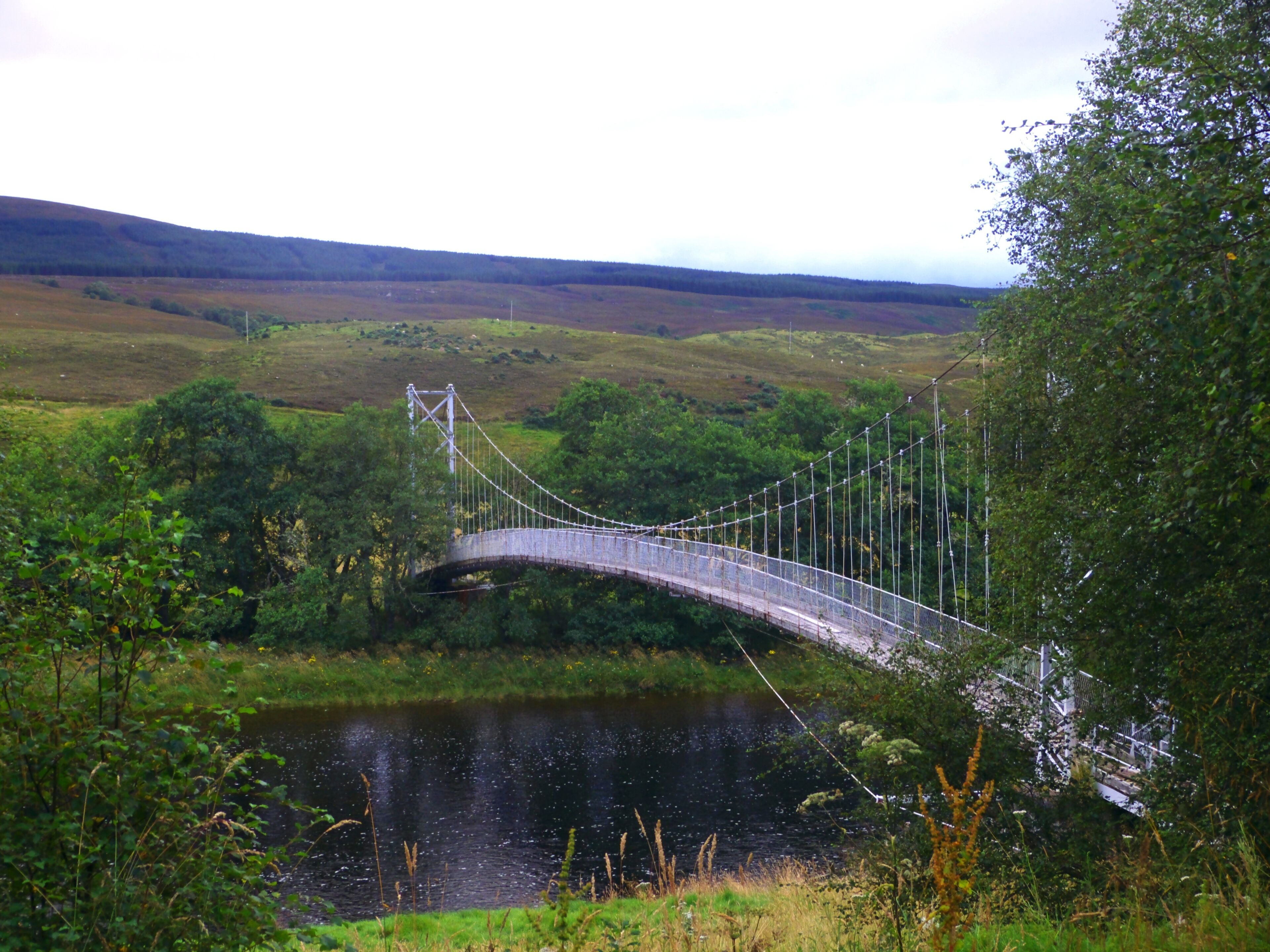 A suspension bridge over a river, surrounded by lush greenery and hills.