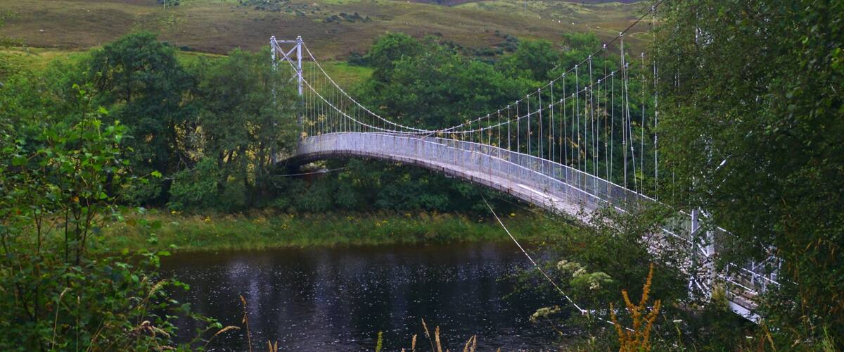 A suspension bridge over a river, surrounded by lush greenery and hills.
