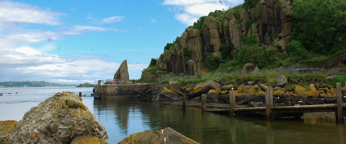 A coastal landscape with a prominent rock formation, a wooden pier, and a clear sky.
