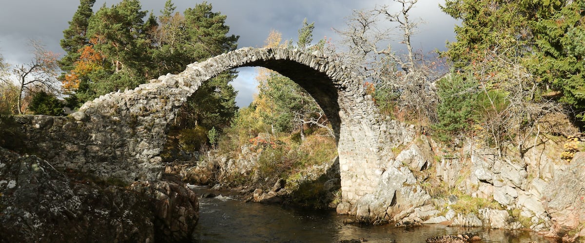 A stone bridge arching over a river, surrounded by trees and rocky terrain.