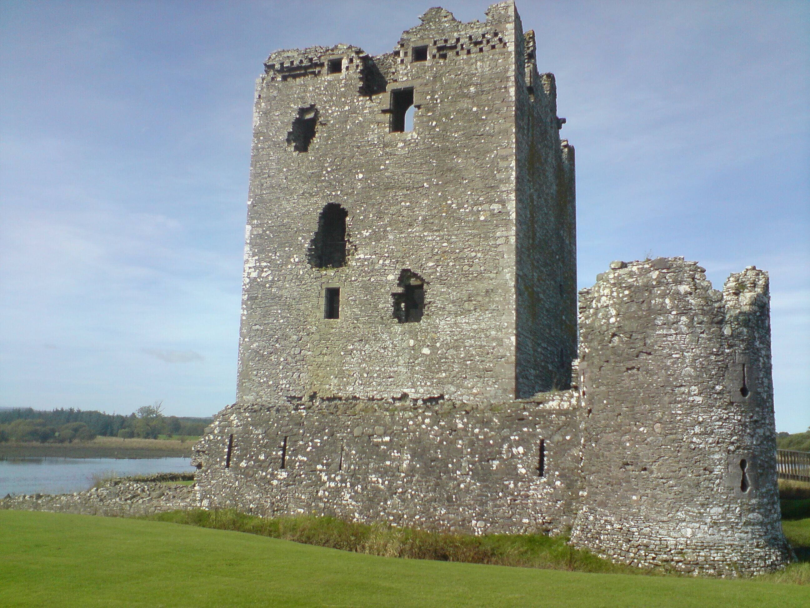 A medieval stone castle with a large tower and a smaller adjacent structure, surrounded by a grassy field and a body of water in the background.