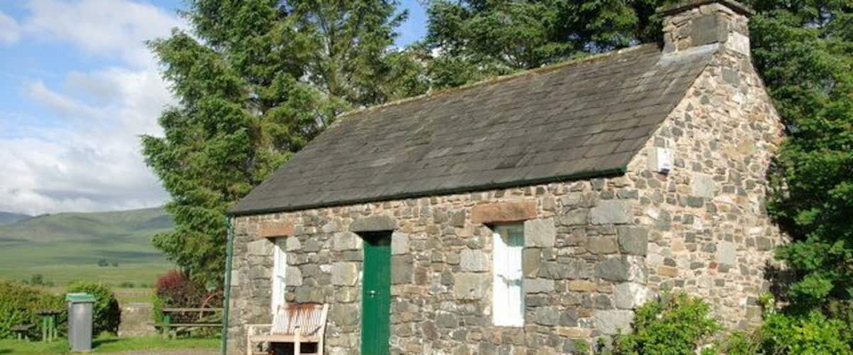 A stone cottage with a green door, surrounded by trees and a grassy field.