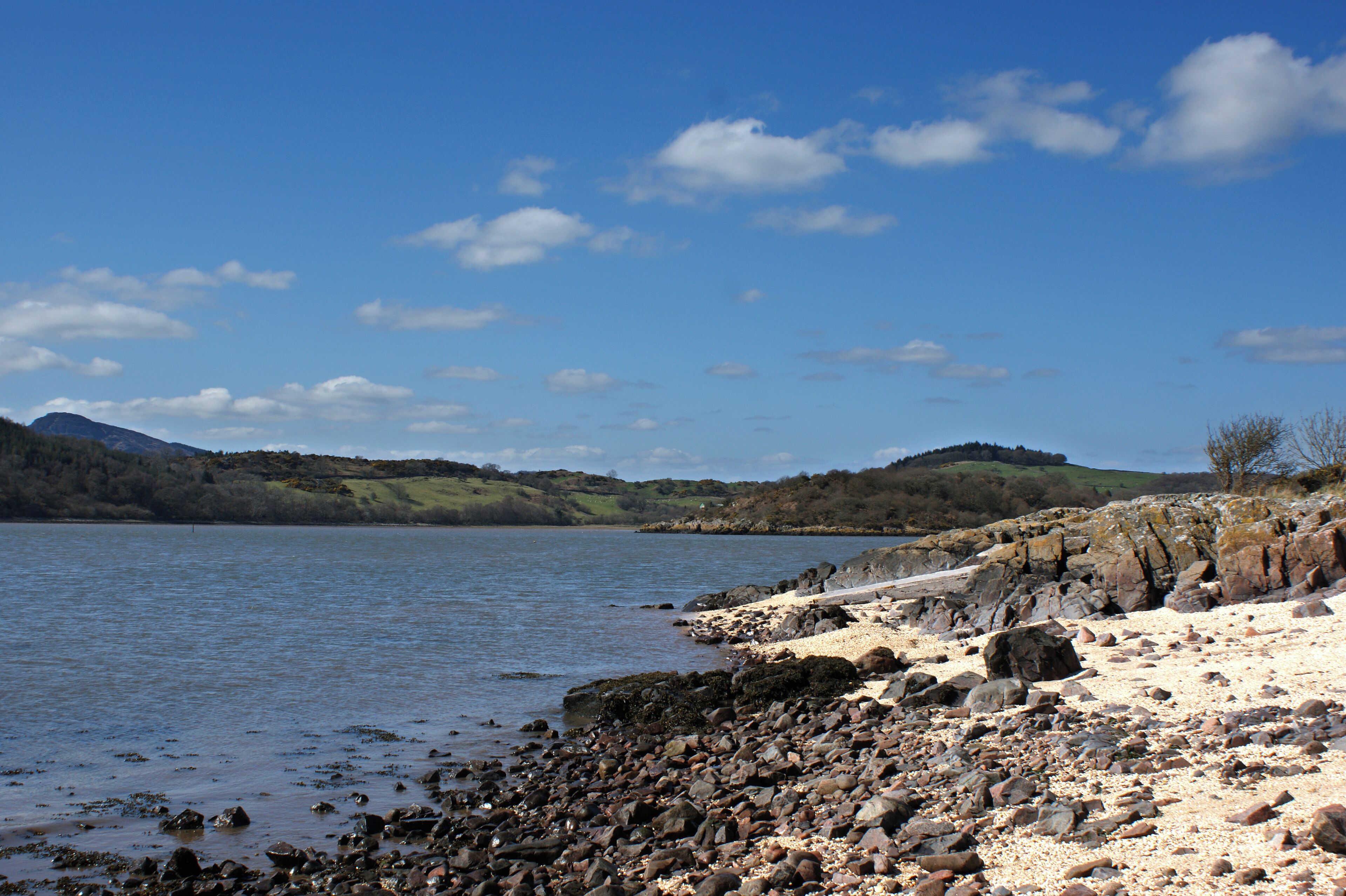 A rocky shoreline with a calm lake, distant hills, and a clear sky.