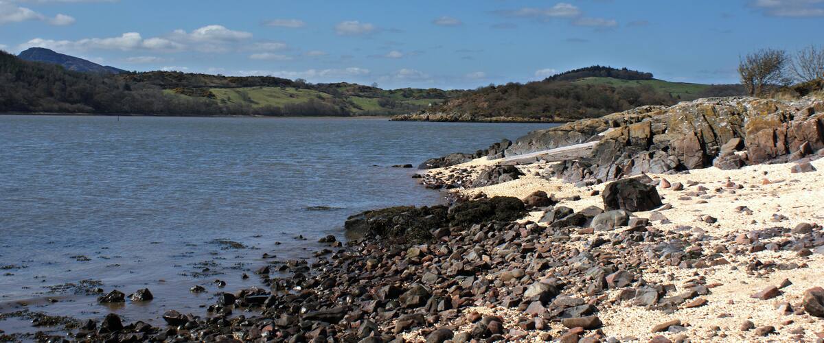 A rocky shoreline with a calm lake, distant hills, and a clear sky.