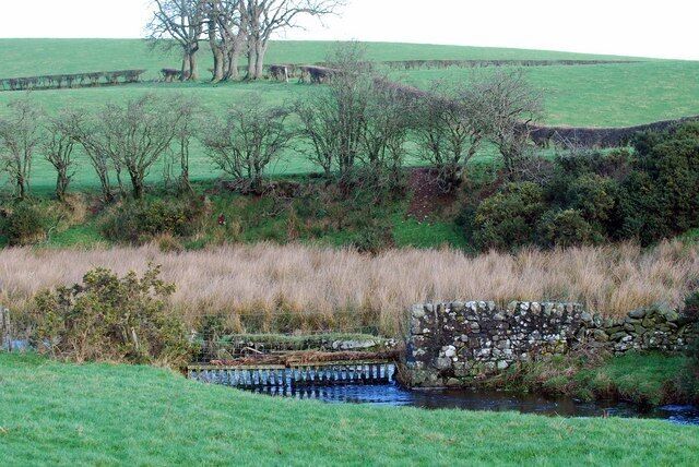 A stone bridge over a stream in a rural landscape with fields and trees.