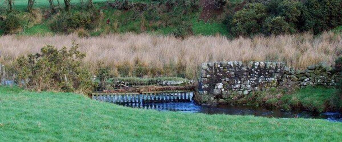 A stone bridge over a stream in a rural landscape with fields and trees.