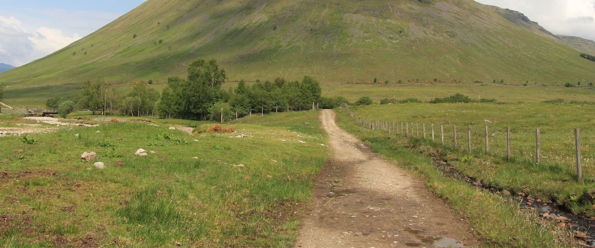 A mountain with a dirt path leading to its base, surrounded by green fields and a fence.