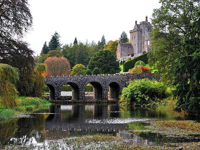A stone bridge over a calm river, surrounded by lush greenery and a castle in the background.