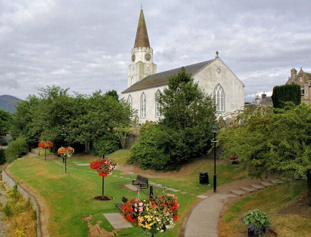 A church with a tall steeple, surrounded by a well-maintained garden with flower beds and a pathway leading to a bench.