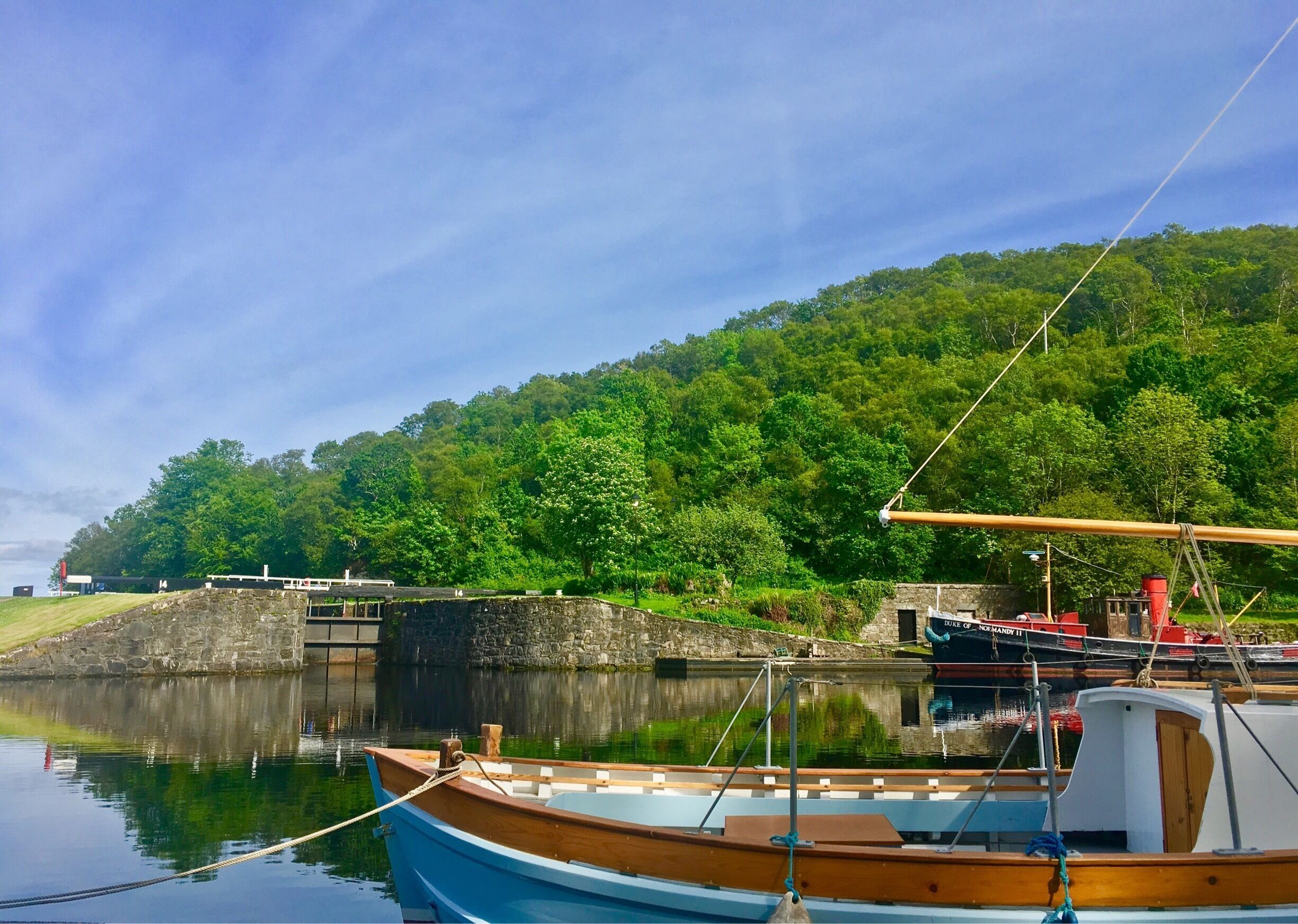 A calm lake with a wooden boat docked in the foreground, a larger boat named "Nor Hardy II" in the background, and a lush green hill with trees.