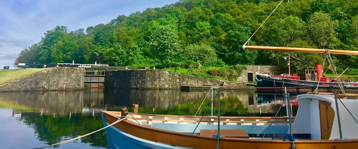 A calm lake with a wooden boat docked in the foreground, a larger boat named "Nor Hardy II" in the background, and a lush green hill with trees.