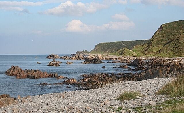A rocky coastline with scattered boulders in the water and a grassy hill in the background.