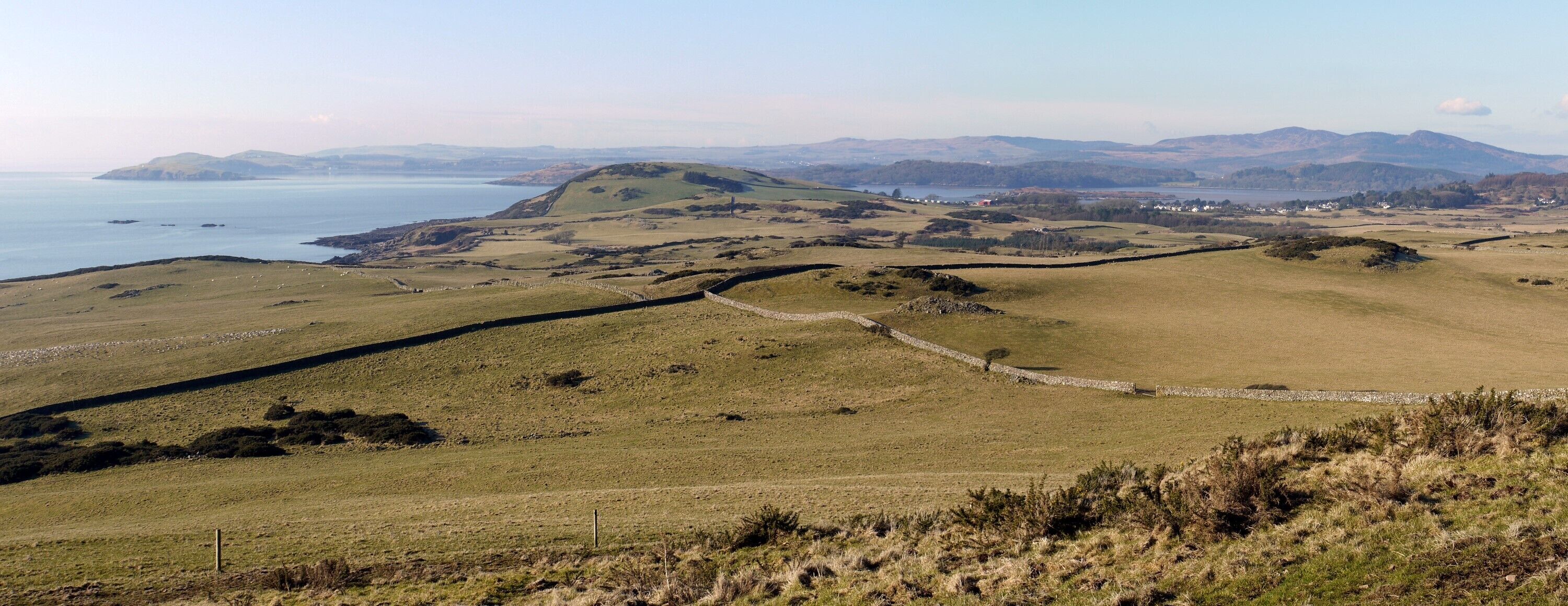 A coastal landscape with rolling hills, a stone wall, and a distant view of the sea and mountains.